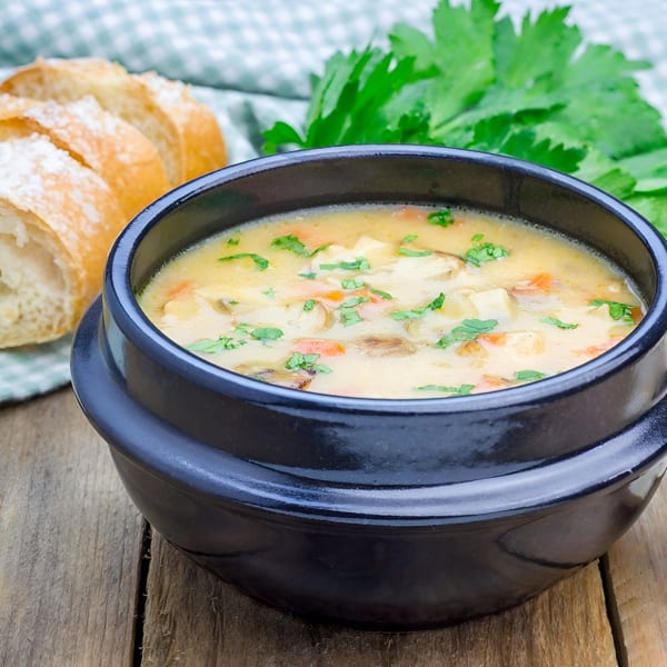 Creamy mushroom and chicken soup in a black bowl, with sliced baguette and parsley on a wooden table.